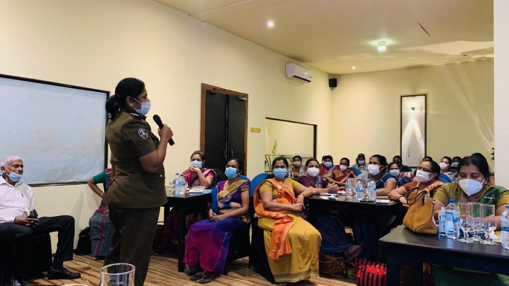 Kvinnor klädda i sarees lyssnar på en kvinna klädd i uniform som pratar i en mikrofon