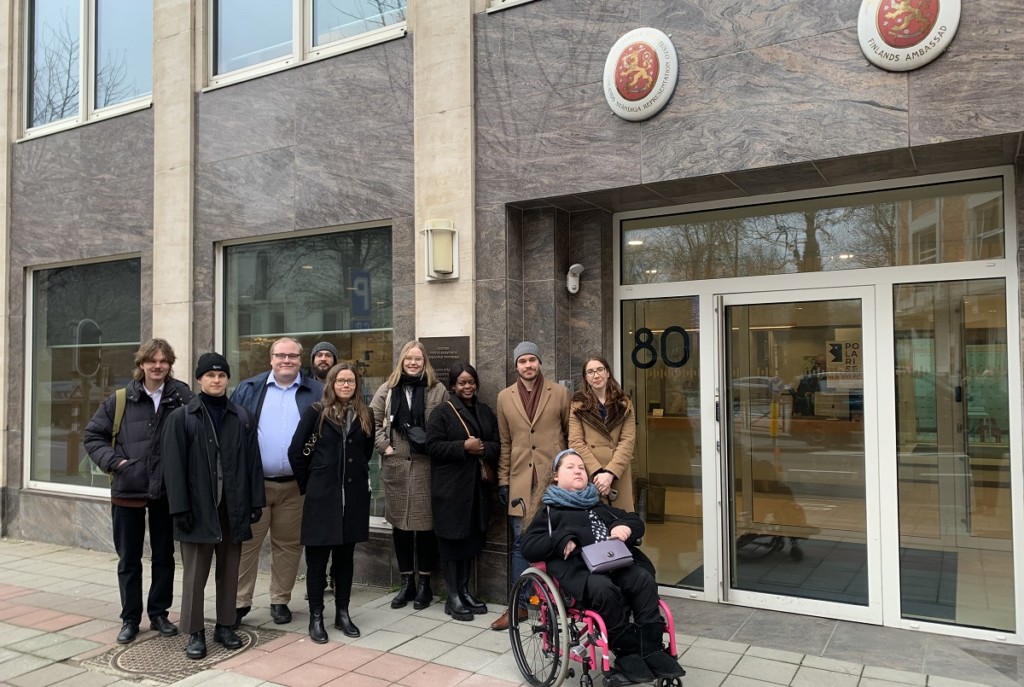 People in a group photo in front of a building that has a sign of Finland's embassy on the wall