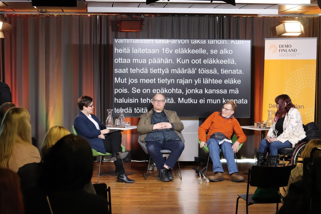 Neljä henkilöä istumassa yleisön edessä, taustalla tekstitystulkkauksen tekstiä isolla ruudulla