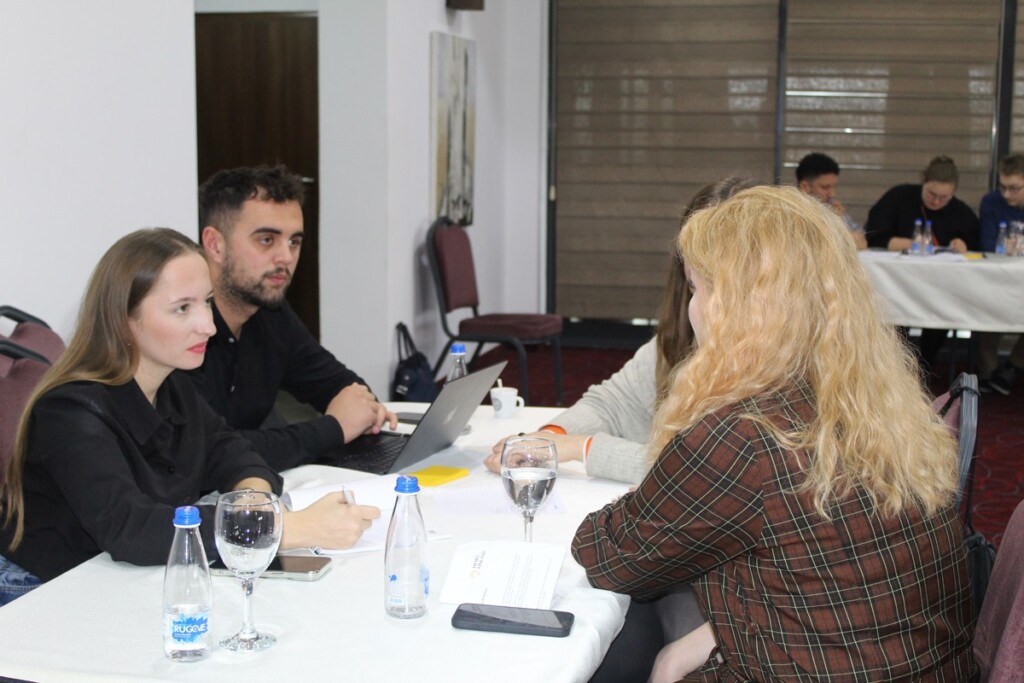 Four people discussing around a table, in the background there is another table with people around it