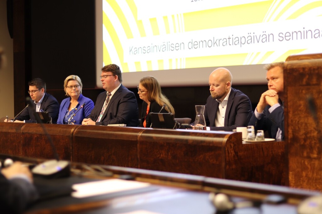 Six people sitting behind a table in a row with microphones in front of them