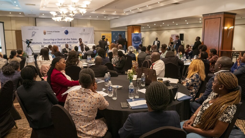 Lots of people, most of whom are Africans, sitting around tables in a room and listening to a woman speaking in front of them