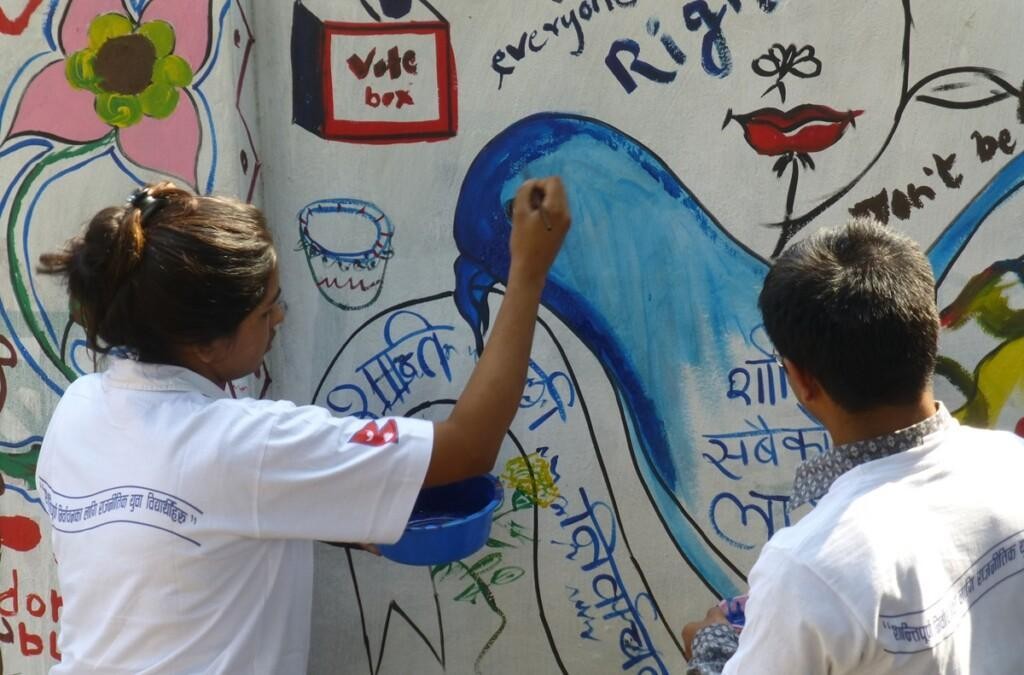 Two people are painting a mural with pictures related to peace and elections and text in the Nepali language.