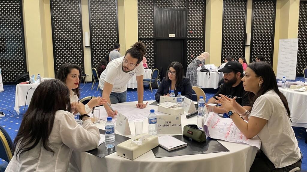 Young people sitting around a table and discussing, one of them is standing and writes on a flipchart.